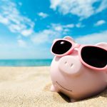 Ein Sparschwein liegt mit einer Sonnenbrille am Strand