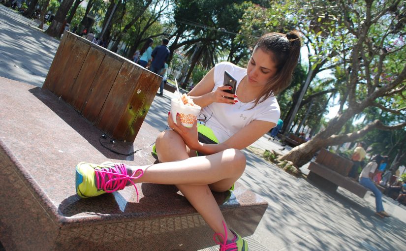Weiblicher Teenager mit Eis und Smartphone auf einer Bank im Park